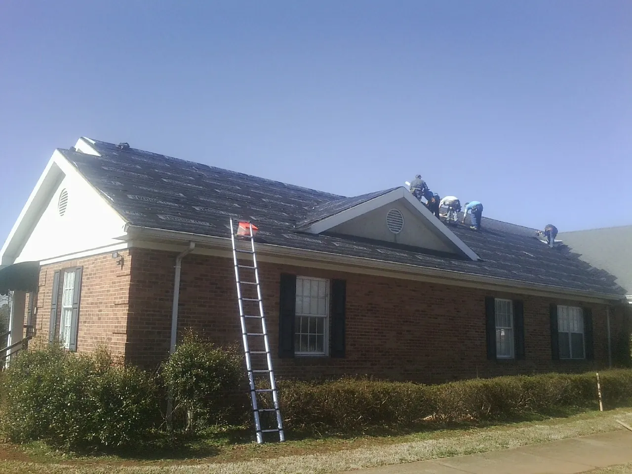 Crew tearing shingles off a brick ranch roof with a ladder set — Metal Roof Repair work by our crew in Pleasant Lake