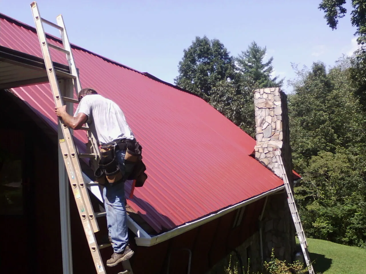 Roofer on a ladder beside a red metal roof and stone chimney — Soffit & Fascia Repair by Roofing Brothers in Pleasant Lake